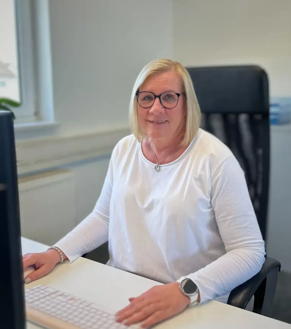 Bild von Astrid Merten, einer Mitarbeiterin, welche glücklich lächelnd im Büro sitzt.